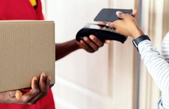 Customer Paying Courier For Delivery Using Smartphone, Receiving Parcel Box At Home. Closeup Of Black Lady Putting Phone To Payment Terminal In Doorway Of Her Home. Mobile Banking Concept. Cropped photo