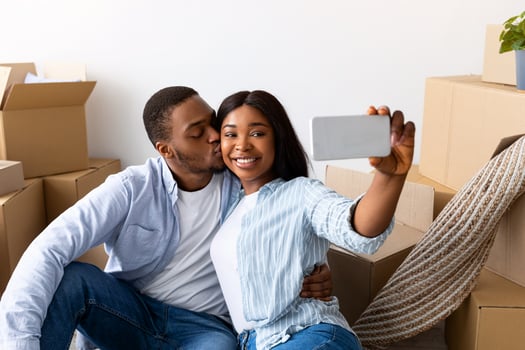 Happy black spouses taking selfie in their new apartment, celebrating moving day, sitting on the floor near carboard boxes. Loving husband kissing wife in cheek photo