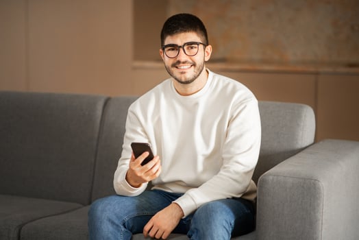 A young Caucasian man with eyeglasses sits comfortably on a gray couch, looking at his smartphone. The cozy home setting reflects a relaxed lifestyle filled with modern decor. photo