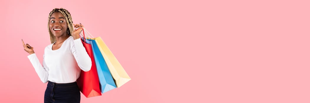 Seasonal Sales And Shopping Concept. Portrait of happy young black woman carrying colorful shopper bags with purchases and gifts, looking back at free space, bright pink studio background, banner photo