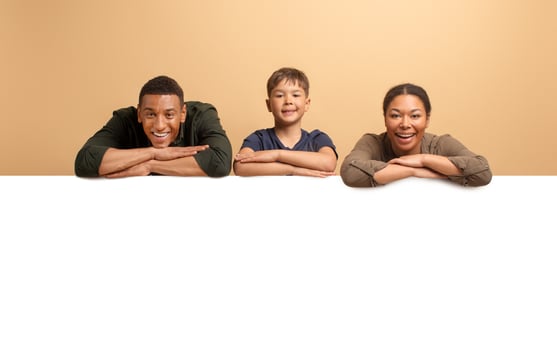 Three family members share a moment of joy, with a father and mother smiling beside their son, leaning on white blank board for advertisement photo