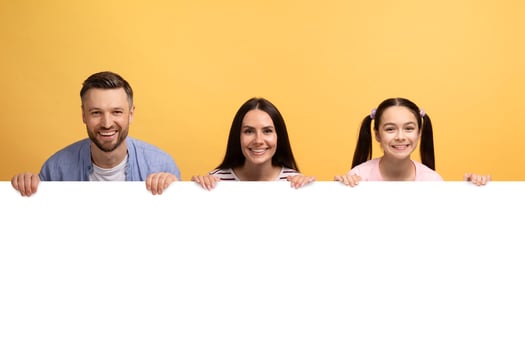 A loving family of three shares a joyful moment against a vibrant yellow backdrop, holding a blank sign. The father, mother, and daughter display warm smiles, embodying affection and connection. photo