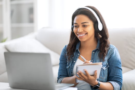 A young black woman smiles while participating in an online learning session from her cozy living room. She takes notes on a notepad, wearing headphones and focused on her laptop. photo
