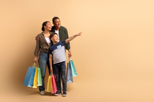 A joyful African American family stands together, holding colorful shopping bags. The teenager points excitedly at copy space, while the parents share smiles, showcasing their bond and happiness. photo