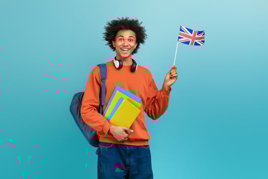 A young African American man with curly hair smiles brightly while holding a small British flag. He wears a casual orange sweatshirt and headphones, carrying yellow and blue folders. photo