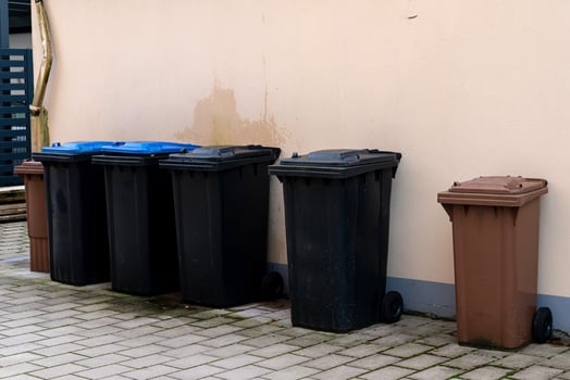 Various trash bins in different colors are lined up against a building photo