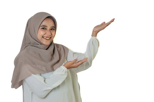 A woman in a beige hijab and white blouse smiles while gesturing with both hands raised, palms up, against a plain white background. photo