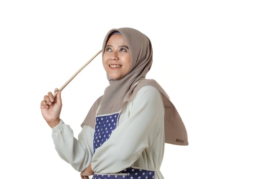 A woman wearing a hijab and a polka dot apron holds a kitchen utensil, smiling confidently, ready for cooking or baking in a bright, positive atmosphere. photo