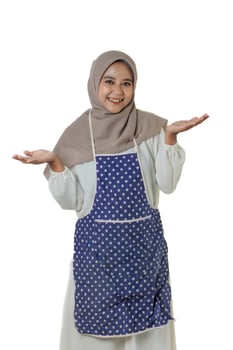 A woman wearing a gray hijab and a polka dot apron smiles and gestures with open hands, suggesting a welcoming or friendly demeanor, ready to help. photo