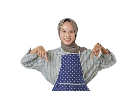 A smiling woman wearing a polka dot apron and a hijab points downward, suggesting a promotion or message below. She stands confidently against a plain background. photo