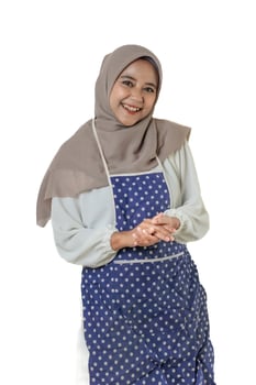 A cheerful woman wearing a beige hijab and a blue polka-dotted apron, smiling warmly at the camera. She exudes positivity and friendliness. photo