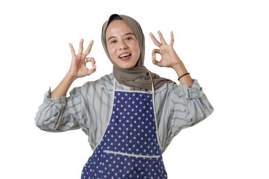 A cheerful woman wearing a blue polka-dot apron and a striped shirt, showing OK hand signs, expressing joy and satisfaction while preparing to cook. photo