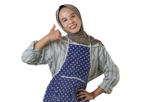 A woman wearing a hijab and a polka-dotted apron poses with a cheerful smile, making a call me hand gesture. She exudes warmth and friendliness. photo