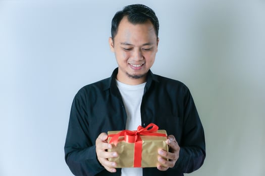 A man wearing a black shirt and a white T-shirt is smiling while holding a gold gift box wrapped with a red ribbon, against a white background. photo