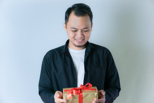 A man wearing a black shirt and a white T-shirt is smiling while holding a gold gift box wrapped with a red ribbon, against a white background. photo