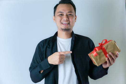 A man wearing a black shirt and a white T-shirt is smiling and pointing to a gold gift box wrapped with a red ribbon, against a white background photo