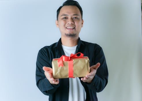 A man wearing a black shirt and a white T-shirt is smiling while presenting a gold gift box wrapped with a red ribbon, against a white background. photo