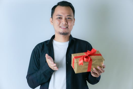 A man wearing a black shirt and a white T-shirt is smiling while presenting a gold gift box wrapped with a red ribbon, against a white background. photo