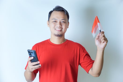 A man wearing a red shirt holds a smartphone and waves an Indonesian flag, showcasing his pride in the concept of celebrating Indonesia's Independence Day. photo