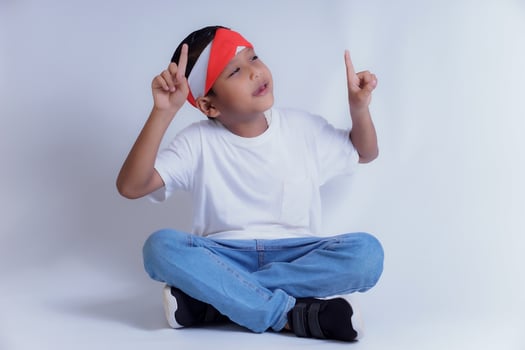 A boy wearing a white shirt and jeans sits cross-legged, pointing up with both hands, smiling brightly. photo