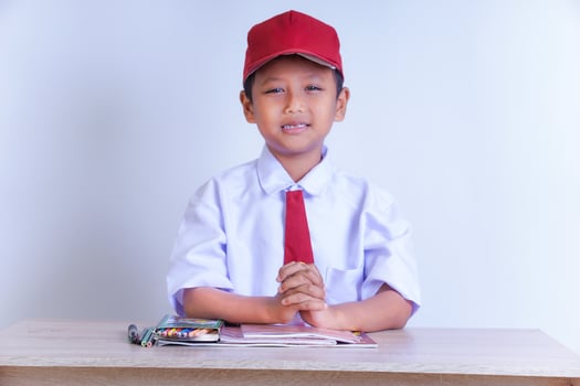 An Indonesian boy in school uniform sits at a table, wearing a white shirt, red tie and red hat. He is smiling with his hands clenched. School supplies sit on the table. The plain background. photo