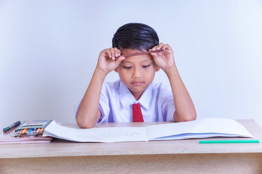 A Indonesian young boy in a school uniform looks contemplative with a pencil on his forehead, sitting at a desk with books and crayons. photo