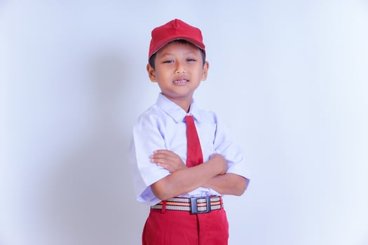 A Indonesian young boy in a school uniform, with a red cap and tie, stands smiling confidently with arms crossed on a white background. photo