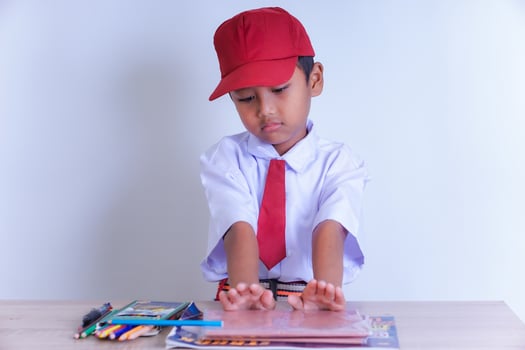 A Indonesian young boy in a school uniform sits at a desk, with a red cap and tie, looking at books and colorful pencils on the table. photo