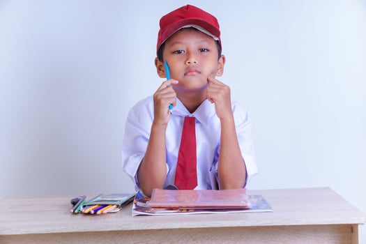 A Indonesian young boy in a school uniform sits thoughtfully with a pen, surrounded by study materials on a desk. photo