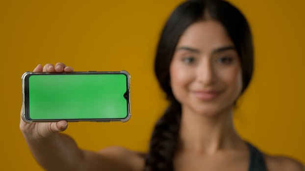 Close up Indian ethnic smiling woman female girl in yellow studio holding phone horizontal showing green empty mobile screen of smartphone chroma key display for advertisement online add offer in cell photo