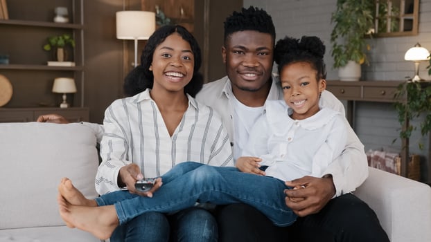 Carefree african american father, mather relaxing with biracial daughter child girl at hme holidays sitting on sofa at home watching television together. Cheerful family relaxing watching tv movie photo