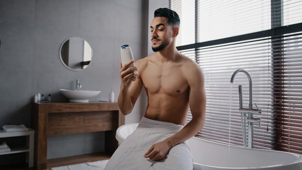 Arab Indian guy muscular naked sexy unshaven man wears white bath towel on hips sits in bathroom getting ready for bathing washing sniffing shower gel morning refreshment with natural male cosmetics photo