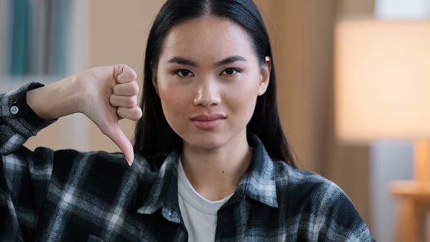 Close up asian millennial woman korean girl wear casual plaid shirt looking at camera showing dislike thumb down disagreement disgust rejection sign bad attitude gesture refusal failure symbol indoors photo