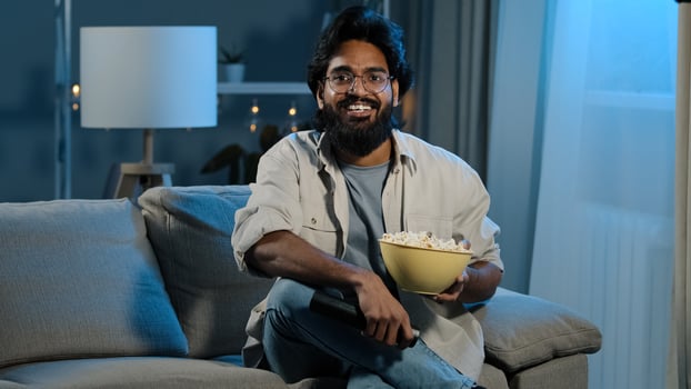 Arab adult bearded man in glasses indian male bachelor after work sits on sofa in dark living room turns on TV with remote control choosing channel watching online movie film program eating popcorn photo