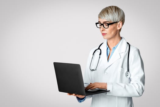 Modern doctor appointment concept. Woman doctor in white coat and glasses writing medical history in laptop isolated on light background, studio shot photo