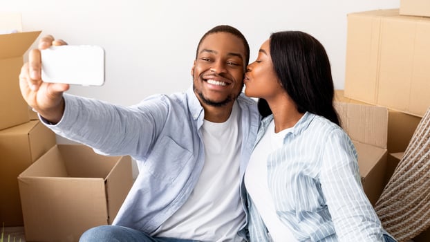 Loving black spouses moving to their own home, taking selfie among carton boxes, wife kissing husband in cheek. Happy young couple making photo of first day in new apartment