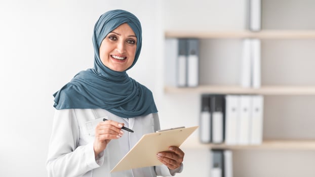 Happy Muslim Female Doctor Posing Smiling To Camera Holding Medical Case Record Standing In Modern Clinic Office Indoors. Portrait Of Mature Middle-Eastern Professional Therapist Lady Wearing Hijab photo