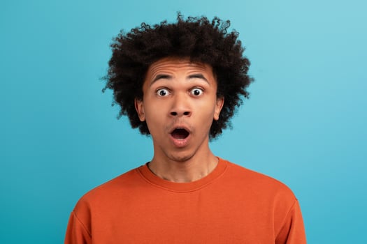 A young African American man with curly hair shows an expression of surprise and shock. He stands isolated against a solid blue background, emphasizing his vibrant reactions. photo