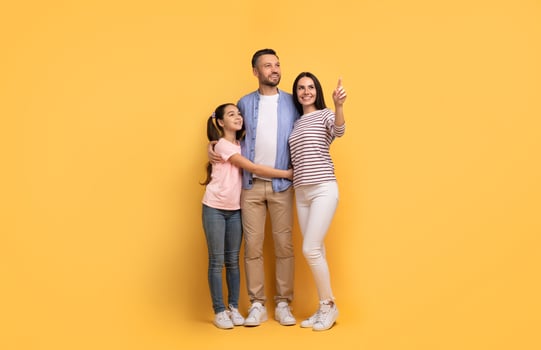 A European family stands together in front of a yellow backdrop, with the daughter hugging her father. The mother points out something, reflecting joy and connection among them. photo