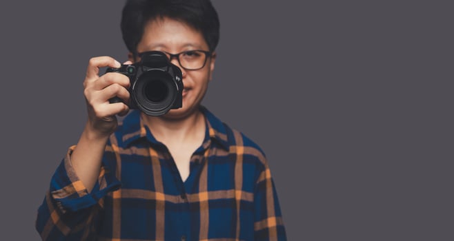 Portrait of a photographer in a blue shirt holding a digital camera while standing on a gray background photo