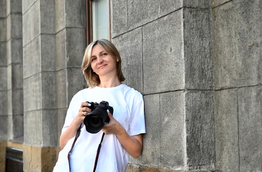 This is a portrait of a woman holding a DSLR camera with a stone wall of a building as a backdrop. She is looking to her left and has a soft smile. photo