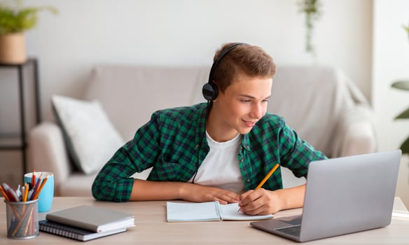 Joyful teen guy writing at notebook and looking at laptop screen, having lesson online, home interior, free space photo