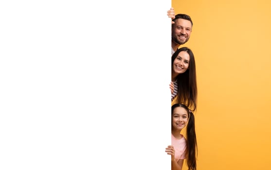 A cheerful family of four stands together, playfully peeking around a large blank surface. Their smiles show affection and happiness in a vibrant yellow background, creating a joyful atmosphere. photo