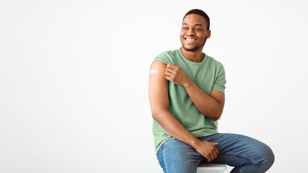 Covid-19 Vaccination. Joyful African Man Showing Vaccinated Arm With Medical Plaster After Coronavirus Vaccine Injection Sitting Over White Studio Background. Covid Immunization. Panorama, Copy Space photo