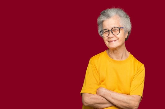 An elderly Asian woman with short gray hair looking at the camera with a smile while standing arms crossed on a red background photo