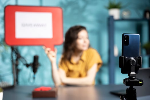Focus on phone used by social media influencer to film giveaway, holding gift box and cardboard sign. Close up of smartphone used by woman recording contest, advertising with copy space board photo