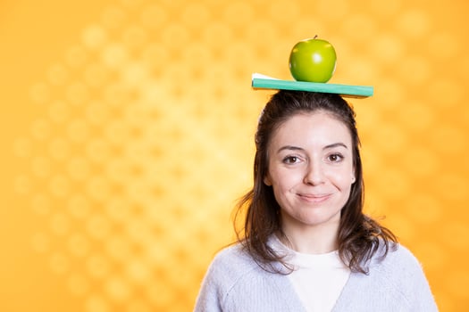 Portrait of jolly woman placing book on head and apple, acting quirky, enjoying reading hobby. Funny geek having fun wearing novel and fruit as hat, toying around, isolated over studio background photo