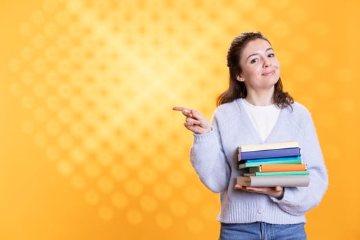 Portrait of smiling woman pointing towards empty copy space while holding pile of books. Radiant person with stack of novels in arms doing advertisement for reading activity, studio background photo