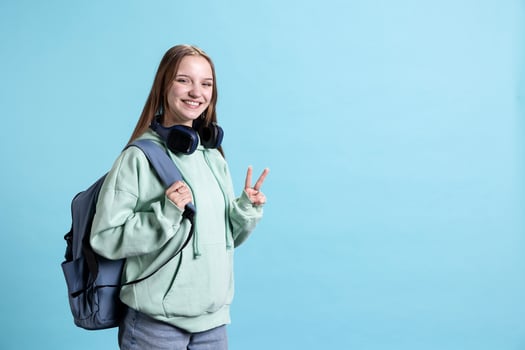 Portrait of happy young woman smiling, doing victory hand sign. Cheerful teenage girl with positive mood showing peace symbol gesturing, isolated over blue studio background photo
