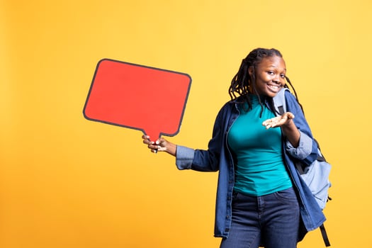 Cheerful woman using red speech bubble sign with empty copy space as advertisement. Upbeat student with backpack presenting thought bubble cardboard used as promotion concept, studio background photo